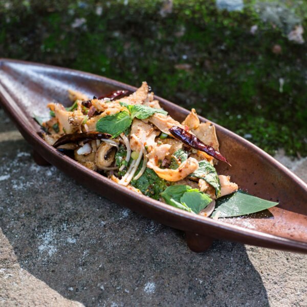 A ceramic dish shaped like a canoe holds a mixed salad with vegetables, garnished with fresh herbs and dried chili peppers. The dish is placed on a rough, stone surface with moss in the background. Sunlight accentuates the colors and textures of the food, making it a Thailand Exclusive Experience at Soneva.