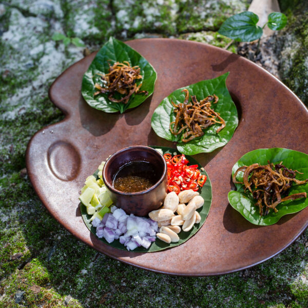 A beautifully presented dish on a rustic wooden plate includes four betel leaves filled with seasoned dried anchovies. Accompaniments of chilies, peanuts, onions, and lime pieces surround a small bowl of dipping sauce. This Thailand exclusive experience is set against a moss-covered stone, embodying the essence of Soneva's luxury dining.