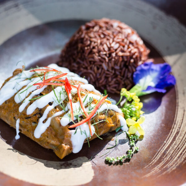 A beautifully plated dish featuring a tamale topped with white sauce, garnished with red chili strips and herbs, accompanied by a mound of brown rice and a single blue edible flower on the side—truly a Thailand Exclusive Experience at Soneva.