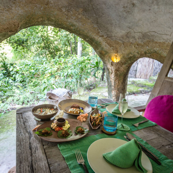 A rustic dining table set with a variety of gourmet dishes and drinks, covered by a thatched wooden roof with arched openings at Soneva Kiri Resort. The lush green forest outside is visible through the arches, creating a serene and natural ambiance. A purple cushion adorns one chair.