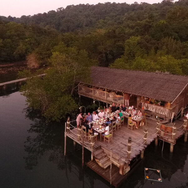 A rustic wooden restaurant built on stilts over water, surrounded by lush green trees. People are dining at outdoor tables under a thatched roof, enjoying the serene, natural setting. A long wooden bridge connects this Thailand exclusive experience to the forest-covered land.