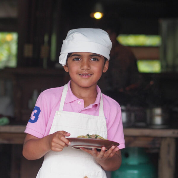 A young child wearing a white chef&#039;s hat, white apron, and pink shirt holds a plate with food, smiling. The background is a dimly lit kitchen with blurred details, hinting at the warmth of a Thailand Exclusive Experience at Soneva, where culinary dreams come to life.