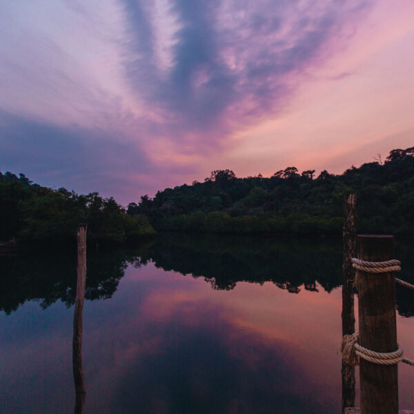 A serene lake at sunset, surrounded by lush, forested hills. The sky is painted in shades of purple and orange, reflecting on the calm water. Wooden posts with ropes jut into the lake, providing a tranquil, picturesque scene—a Thailand Exclusive Experience reminiscent of Soneva resorts.