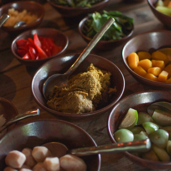 A variety of colorful ingredients are displayed in brown bowls on a wooden surface, embodying a Thailand exclusive experience. Each bowl contains a different food item, such as diced vegetables, red chilies, spices, and what appears to be a paste or sauce. Spoons are placed in several of the bowls.