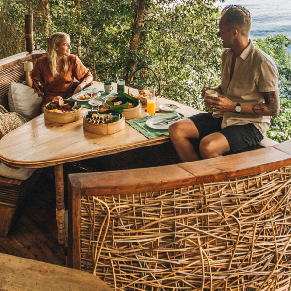 Two people enjoy a meal in a treehouse restaurant with woven wicker seating. The setting, part of the Thailand Exclusive Experience by Soneva, is surrounded by lush greenery, overlooking a scenic ocean view. Various dishes and drinks are spread out on the wooden table. The ambiance is serene and nature-filled.