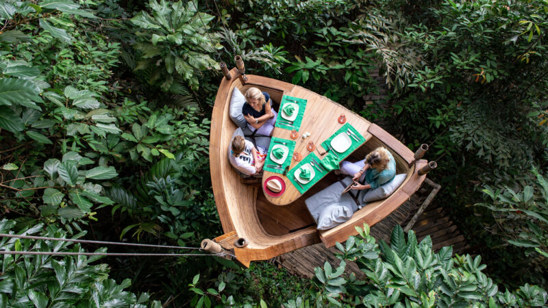 Three people dining at a wooden table shaped like a boat, suspended high among dense green foliage. The table is set with plates, glasses, and napkins. The diners sit on cushioned bench seating, surrounded by lush tropical vegetation—an exclusive Soneva experience in Thailand.