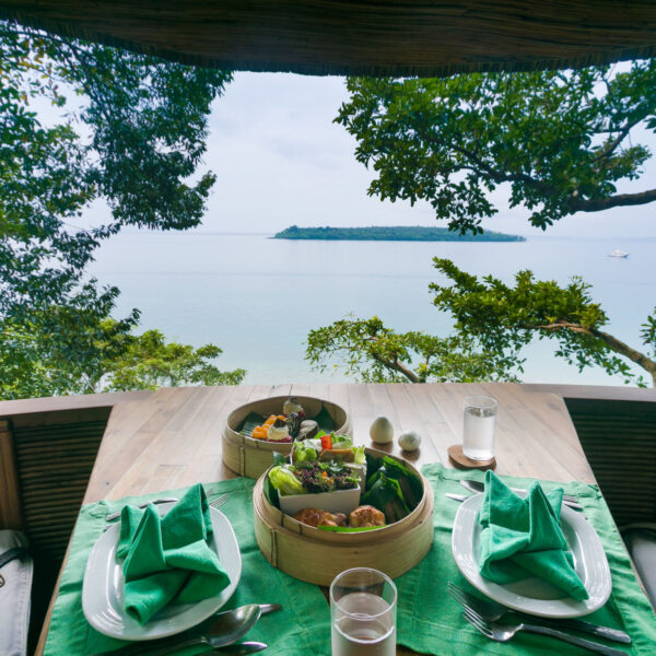 A serene outdoor dining setting with a wooden table covered by a green cloth is adorned with plates of food and bamboo steamers. This Thailand Exclusive Experience at Soneva overlooks a calm, expansive body of water surrounded by lush trees and the silhouette of an island in the distance.