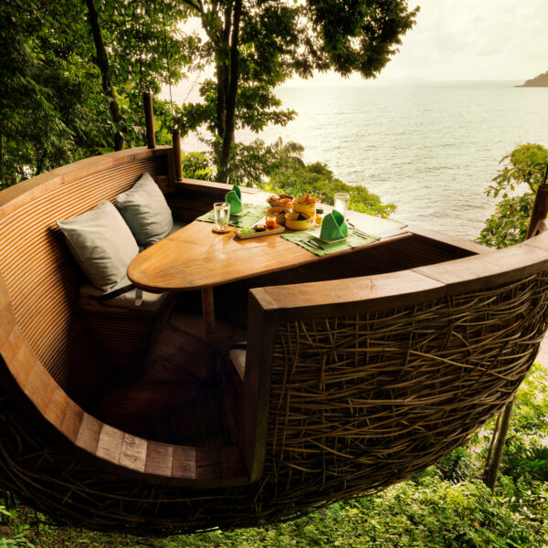 A unique dining setup features a boat-shaped wooden platform suspended in the trees, overlooking a serene body of water. This Thailand exclusive experience at Soneva includes a table with neatly arranged green napkins, glasses, and plates of food surrounded by cozy seating with cushions. Lush greenery surrounds the scene.