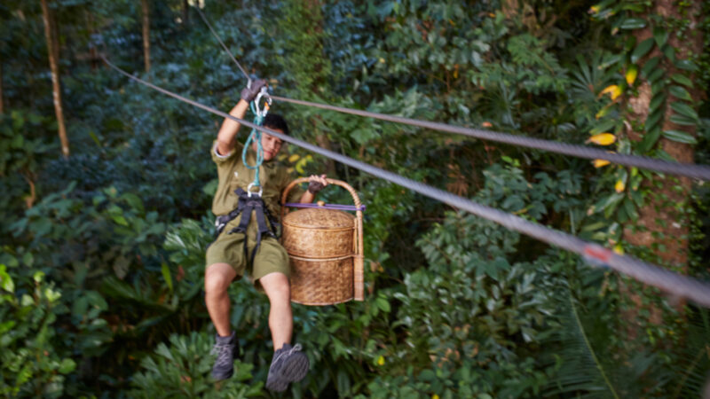 A person in adventure gear rides a zipline through a dense forest, carrying a large woven basket in one hand. The lush foliage below suggests a tropical paradise, making this Thailand exclusive experience truly unforgettable.