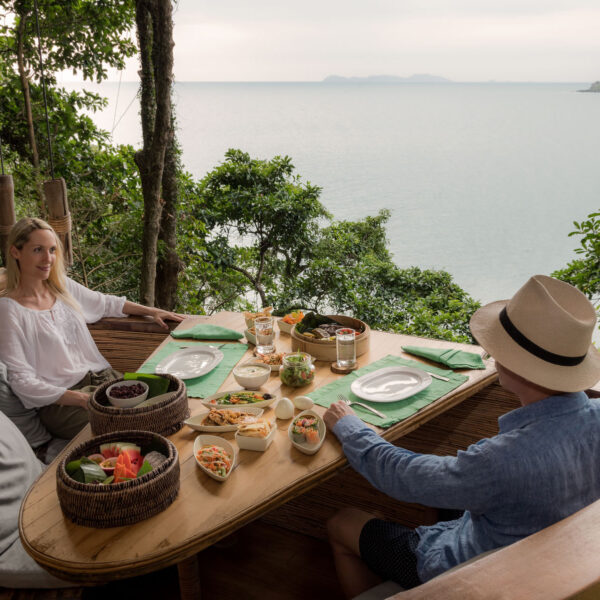 A woman and a man enjoy a meal at a round wooden table set with various dishes, overlooking a lush forest and the serene ocean view of their Thailand Exclusive Experience at Soneva. The woman is reading a menu, while the man wears a hat and looks out towards the water. The atmosphere is peaceful and natural.