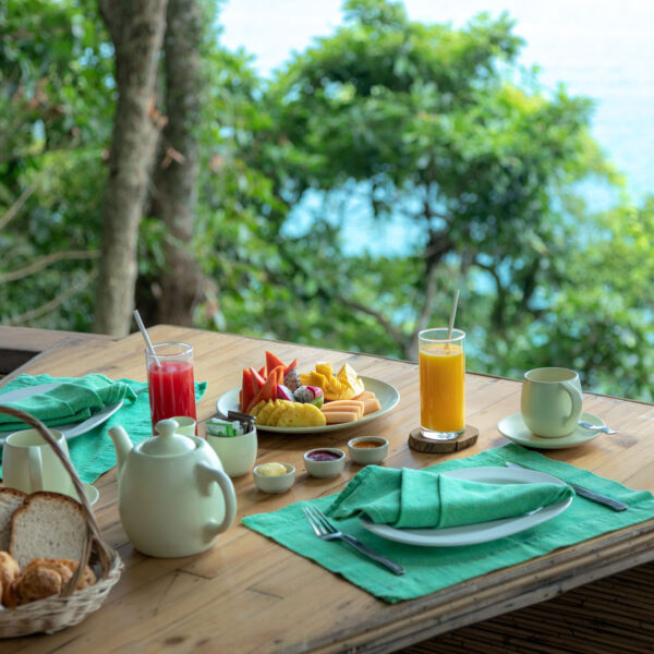 A picturesque outdoor breakfast setting on a wooden table adorned with green napkins. The table holds a basket of bread and pastries, a tea pot, cups, plates of fruit, various small dishes, and glasses of red and orange juice. This Thailand exclusive experience offers lush greenery and an ocean view in the background at Soneva.