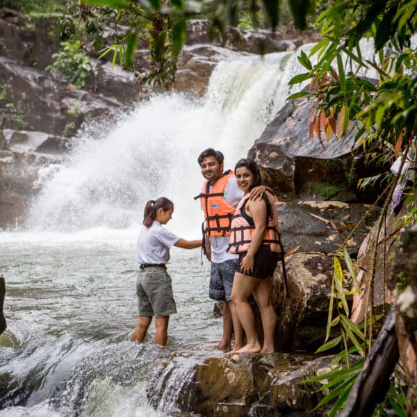 Three people stand near the edge of a rocky waterfall amidst lush greenery. Two of them, partaking in this Thailand exclusive experience, wear orange life vests, while the third person in a white shirt and gray shorts helps. The waterfall cascades behind them with pure Soneva elegance.