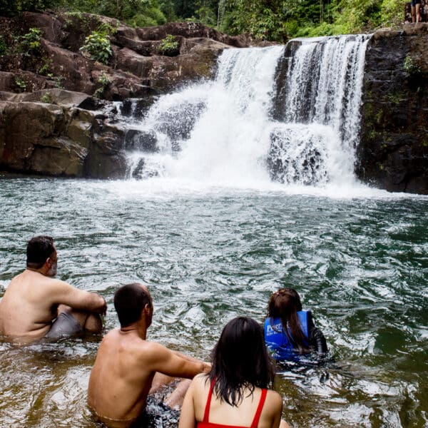 Four people are sitting in a natural pool at the base of a small waterfall surrounded by rocks and lush greenery. Their backs are to the camera as they face the waterfall, with two wearing swimwear and one in a blue shirt—truly an exclusive experience that feels like a hidden gem in Thailand.