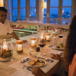 A chef engages in conversation with a woman seated at a candlelit table in a restaurant. The table is set with various plates of Soneva’s exquisite food, and the background shows large windows revealing a twilight sky reminiscent of a Maldives luxury resort.