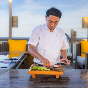 A chef in a white uniform, embodying the essence of Soneva Career, prepares food on a wooden cutting board at an outdoor kitchen with a seaside view. The setup includes some vegetables on the board, a few dishes, and comfortable seating with yellow cushions in the background.