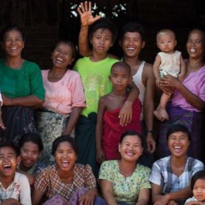 A group of people, including men, women, and children, are gathered in front of a wooden doorway in the Maldives Namoona tradition, smiling and laughing. The group consists of individuals of various ages, with one person holding a baby. An older man wearing a turban stands at the left.