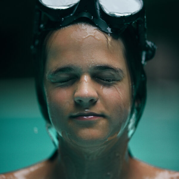 A person with their eyes closed, wet skin, and wearing a black diving mask on their head is partially submerged in water. The image captures the person from the shoulders up against a blurred outdoor background, evoking a serene and peaceful moment reminiscent of Soneva Exclusive Offers.