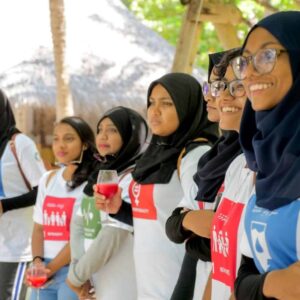 A group of women in headscarves and t-shirts stands together outdoors, smiling and holding drinks. Their t-shirts feature colorful, printed designs that capture the essence of Maldives Namoona. The background includes trees and a thatched-roof structure, suggesting a tropical setting.