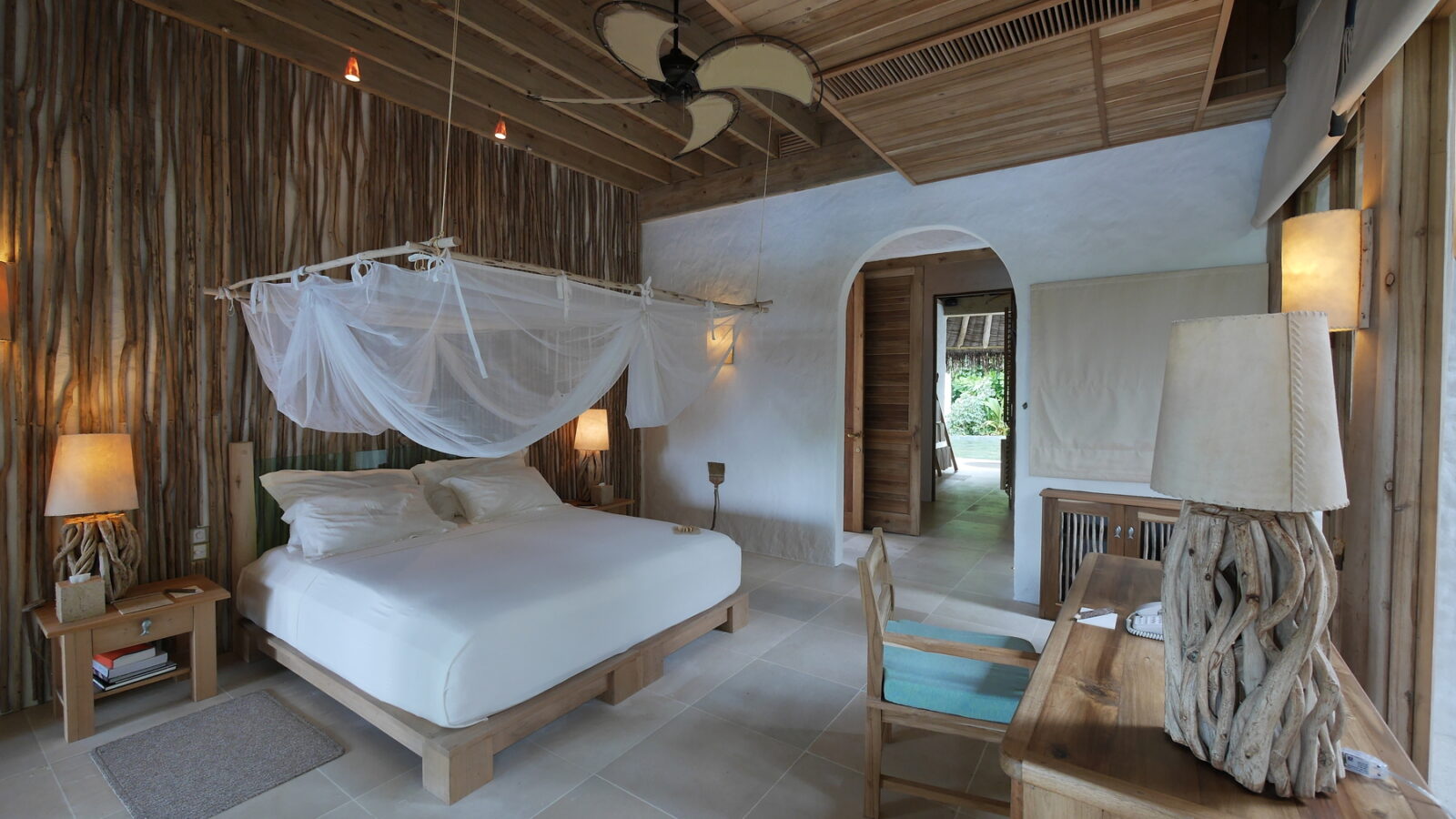 Experience a spacious beach retreat villa, featuring a canopy bed draped in white mosquito netting. The room boasts wooden accents, a ceiling fan, and lamps with natural wood bases. Step through the doorway for a lush outdoor view that captures the essence of the Maldives.