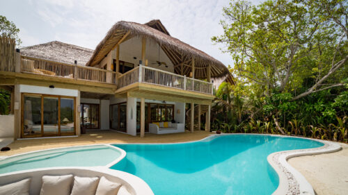 A luxurious Maldives beach retreat villa with a thatched roof and wooden accents, featuring a spacious deck overlooking a curved swimming pool. Surrounded by lush greenery under a partly cloudy sky, the villa boasts outdoor seating and large glass doors.