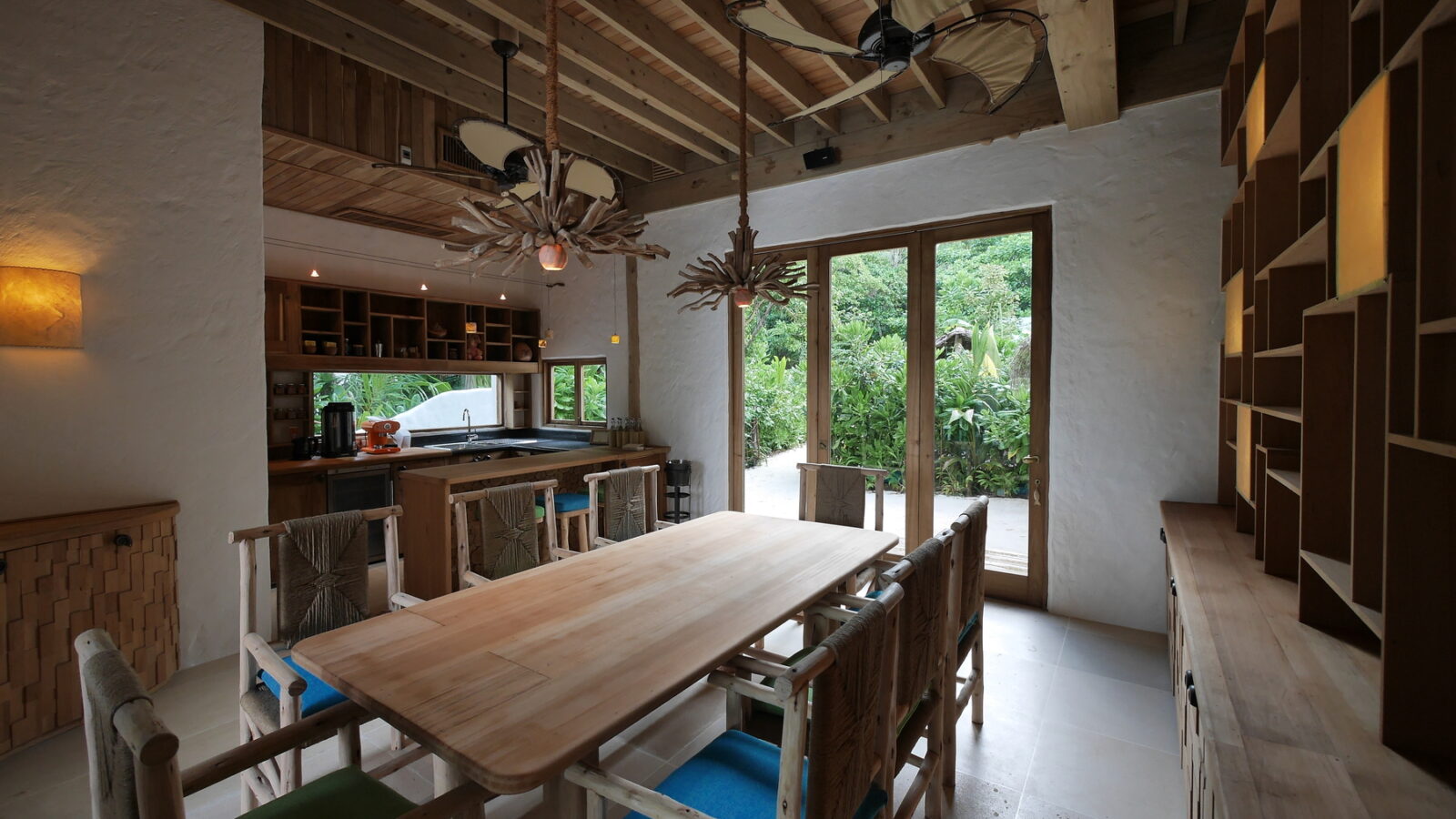 A rustic dining room in a spacious beach retreat villa showcases a wooden table and chairs beneath a ceiling with exposed beams. Large windows reveal a lush garden, reminiscent of the Maldives, while unique light fixtures and open shelves add charming details.