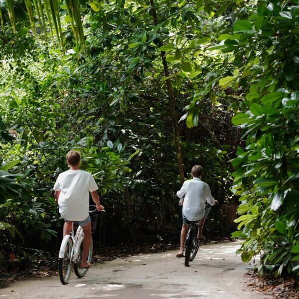 Two people are riding bicycles on a dirt path surrounded by lush, dense greenery. Both cyclists are wearing casual white shirts, and the scene suggests a peaceful, nature-filled environment.