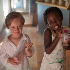 Two young children enjoying ice cream indoors. The child on the left holds a spoon and cup, wearing a white shirt, while the child on the right is smiling, wearing a paint-splattered apron with a cup in hand. They stand on a wooden floor.