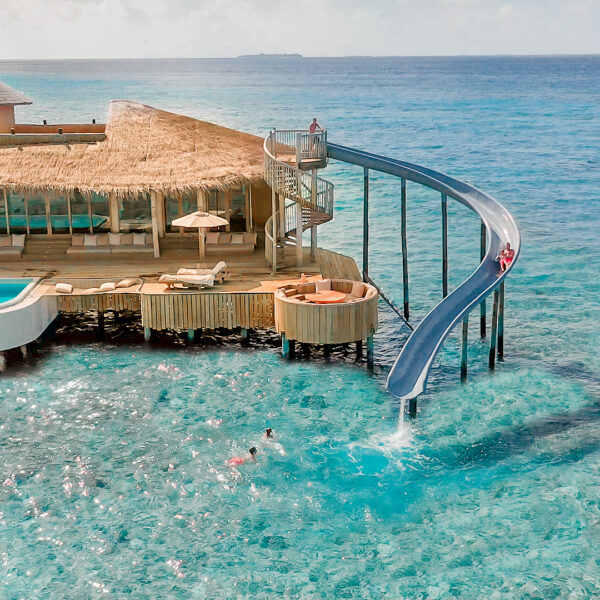 A luxurious overwater villa with a thatched roof features a long slide into the turquoise ocean below. Sun loungers and a pool are on the deck, with people swimming and enjoying the tropical waters.