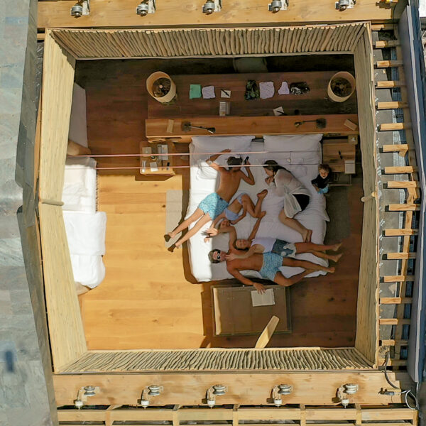 Aerial view of a family of four relaxing on a bed inside a wooden cabin. The room features minimal decor, including two side tables and some papers. The cabin has a rustic and open design with a partial view of the surrounding thatch roof.