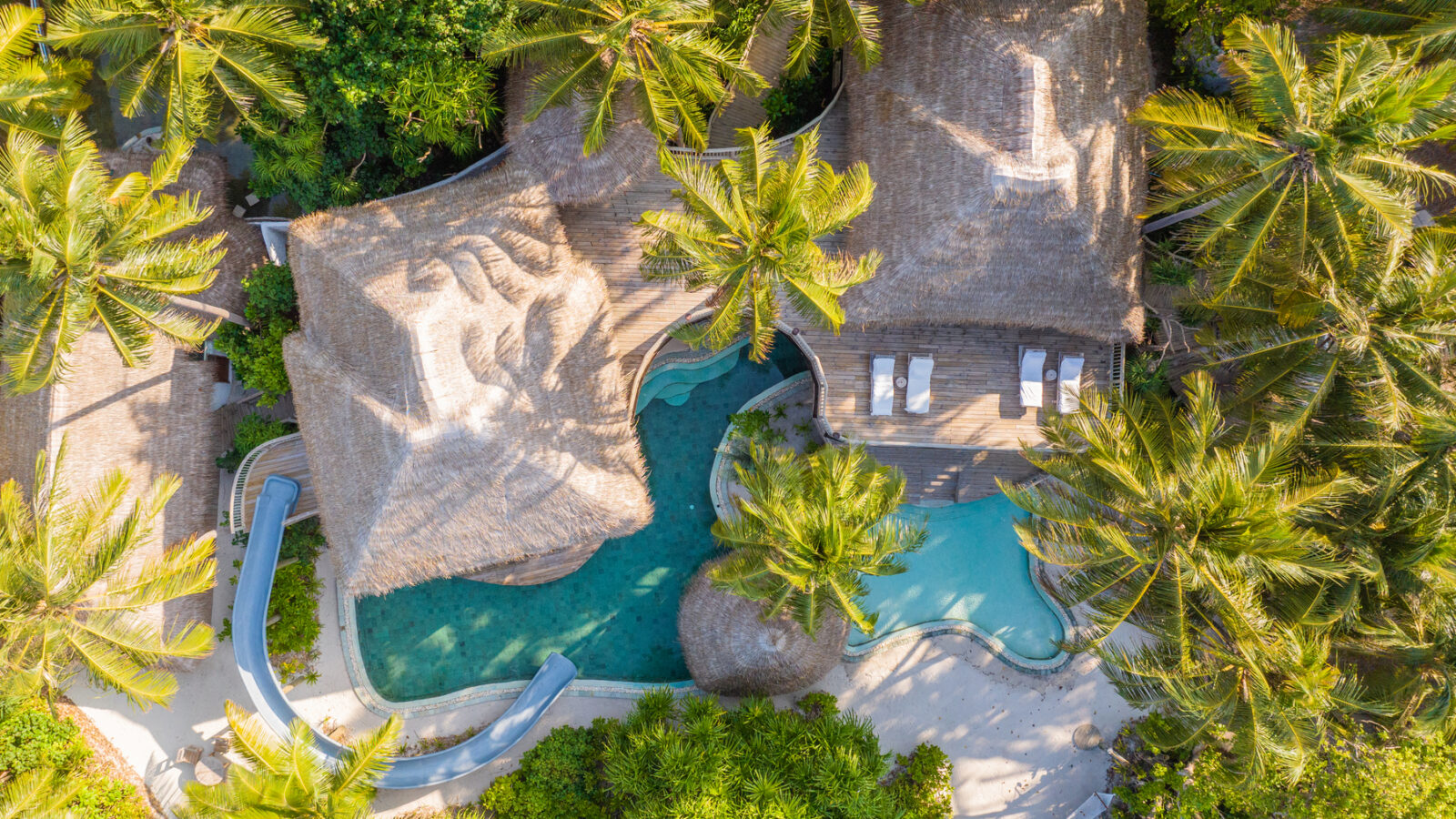 Aerial view of a tropical resort with two thatched-roof huts nestled in a lush jungle reserve. A curvy water slide winds into a turquoise pool. Sun loungers are arranged on the deck near the pool, creating a serene, vacation-like setting amidst verdant palm trees.