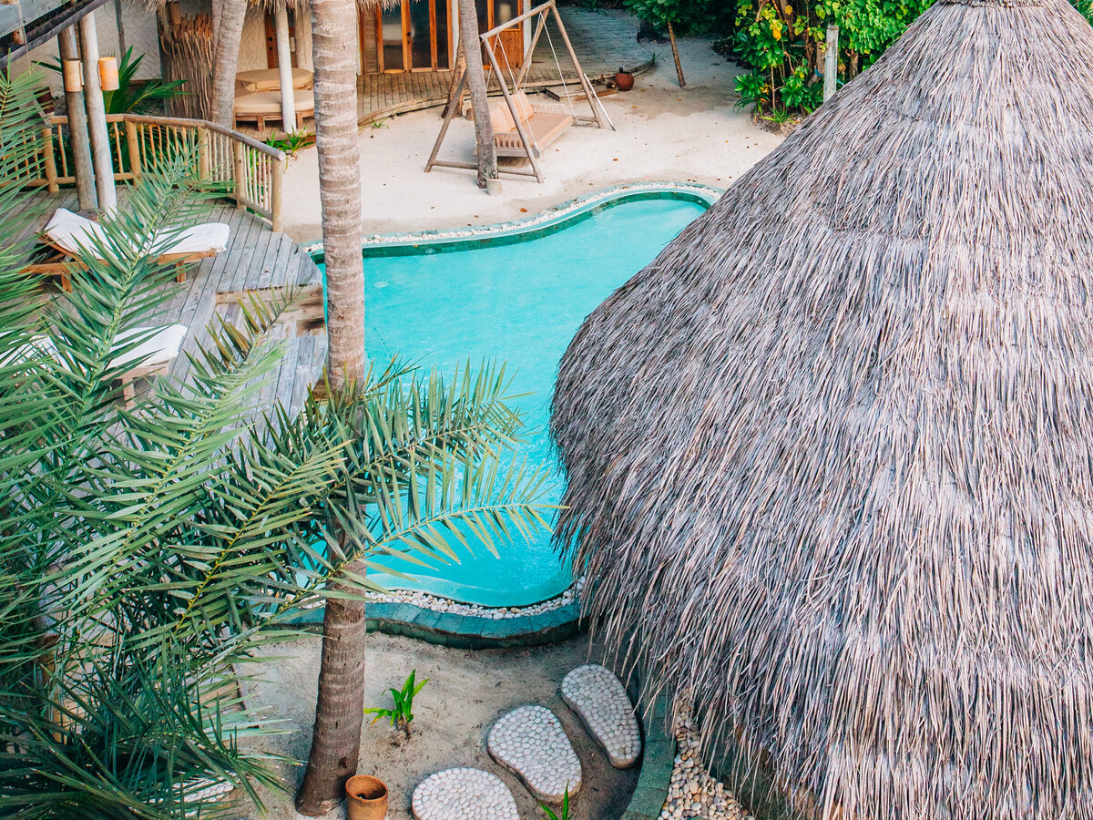 Aerial view of a tropical resort nestled in the Jungle Reserve, featuring a turquoise pool surrounded by lush greenery. Thatched-roof huts and a wooden swing enhance the sandy area. Palm trees and stone pathways add to the serene island ambiance.