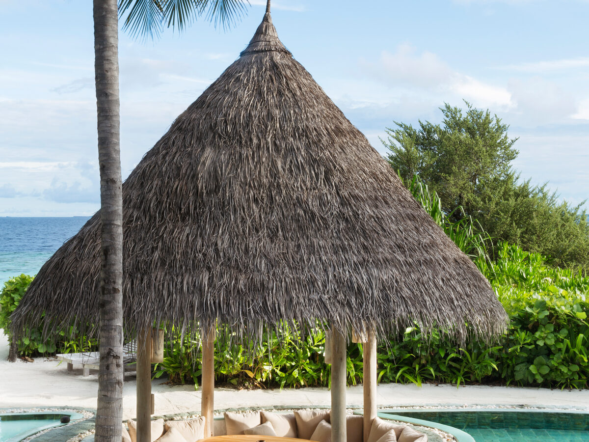 A serene tropical scene with a thatched gazebo, nestled in a jungle reserve, surrounded by lush greenery beside a turquoise pool. Tall palm trees frame the setting, with the ocean visible in the background under a partly cloudy sky.