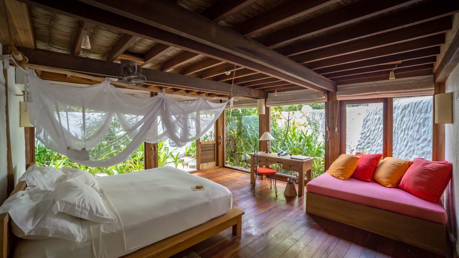 A cozy bedroom with wooden beams and flooring, reminiscent of the charming Soneva Fushi Villas in a Maldives luxury resort. The room features a large bed with white bedding, a wooden desk with a red chair, and a cushioned bench adorned with pink and red pillows. Large windows and a glass door offer lush green garden views outside.