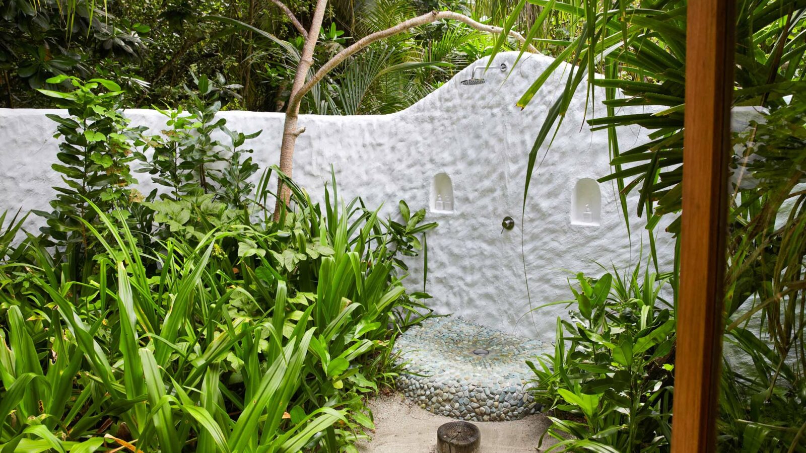 An outdoor shower set within a lush, tropical garden at the Maldives luxury resort, Soneva Fushi Villas. The shower features a white, textured wall with built-in niches and a pebble stone floor. Surrounding the shower are dense green plants and trees, creating a serene and private natural space.