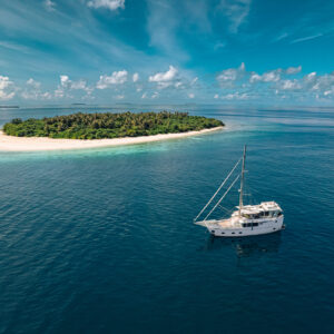 A small, lush island with a sandy beach is surrounded by clear blue water under a partly cloudy sky. A white luxury yacht is anchored nearby in the calm sea, creating a serene Maldives holiday scene.