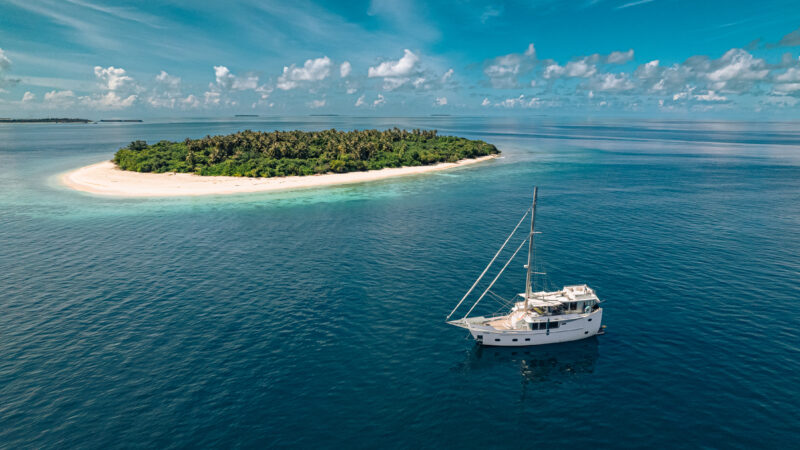 A small, lush island with a sandy beach is surrounded by clear blue water under a partly cloudy sky. A white luxury yacht is anchored nearby in the calm sea, creating a serene Maldives holiday scene.