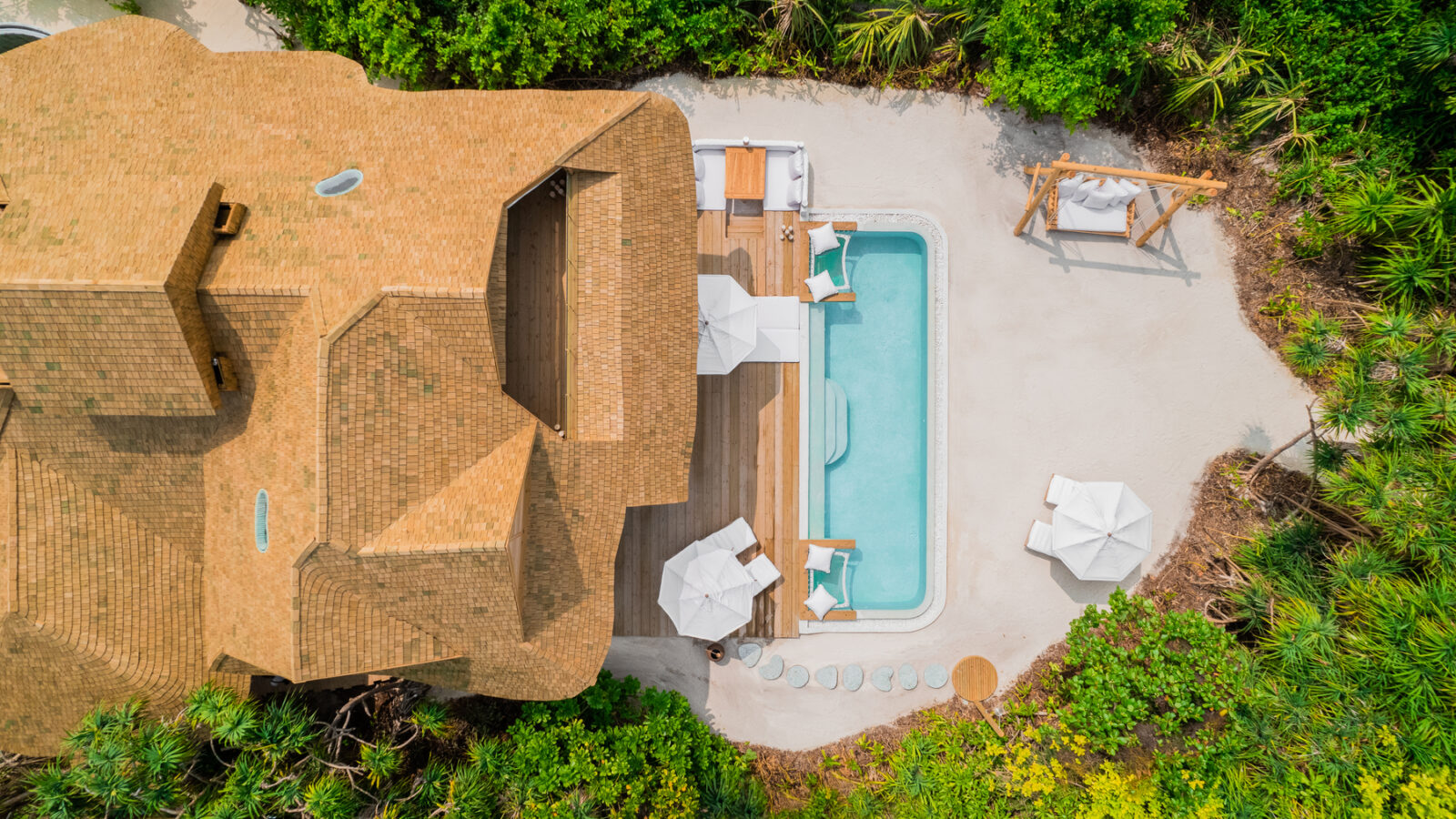 Aerial view of a Soneva Jani Island villa with a pool, enveloped by lush greenery. The patio area features sun loungers with umbrellas and a wooden swing, perfect for a family retreat. The villa's brown roof complements the tropical paradise setting beautifully.