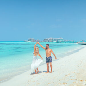 A couple dances joyfully on a pristine, white sandy beach with crystal-clear turquoise water in the background. The woman wears a flowing white dress, and the man is in blue swim trunks. Overwater bungalows, part of the Soneva Exclusive Offers, can be seen in the distance under a bright blue sky.