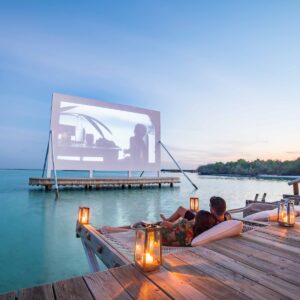 A couple relaxes on a wooden deck over turquoise water, watching a movie projected on a large screen at sunset. Illuminated with lanterns, the cozy, romantic atmosphere near the serene tropical shoreline is part of Soneva's exclusive offers.