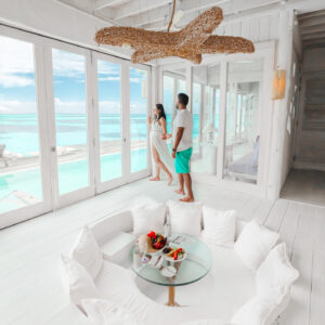 A couple stands by large glass doors, gazing at a turquoise ocean and blue sky. The room is bright with white decor, featuring a sunken seating area with white cushions and a glass table with snacks and drinks. A wooden light fixture hangs from the ceiling above them, accentuating the Soneva Exclusive Offers ambiance.