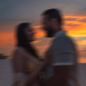 A blurry photo of a couple standing closely together on a beach at sunset. The sky is vividly painted with shades of orange, yellow, and blue. Both individuals are out of focus, adding a dreamy, romantic ambiance to the scene—perfect for capturing those Soneva Exclusive Offers moments.