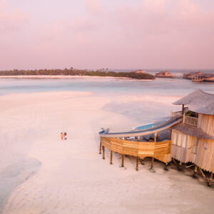 A breathtaking view of a tropical beachfront villa built on wooden stilts with a slide extending into the water. Clear, shallow water and white sandy beach surround the villa. Under a pastel pink and purple sky, two people stroll along the shore, taking advantage of Soneva Exclusive Offers.