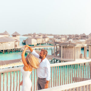 A couple dances on a wooden platform overlooking overwater bungalows in a tropical setting. The woman, in a white dress, spins gracefully while the man, in a white shirt and beige pants, smiles and holds her hand. The turquoise waters and bungalows create a serene backdrop for this Soneva Exclusive Offers moment.