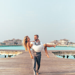 A man in a gray suit is carrying a woman in a white dress along a wooden pier with overwater bungalows in the background. Both are barefoot and smiling, enjoying a sunny day by the turquoise sea, reminiscent of Soneva Exclusive Offers.