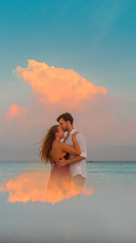 A couple stands waist-deep in calm ocean water, tenderly embracing under a dramatic sky. A large, orange-tinged cloud dominates the background, reflecting on the water's surface. The serene setting evokes a sense of romantic tranquility, reminiscent of Soneva Exclusive Offers' idyllic escapes.