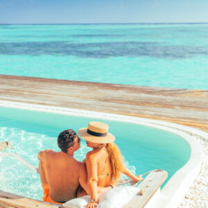 A couple in swimwear sits on the edge of a private pool overlooking a stunning turquoise ocean. The woman wears a wide-brimmed hat and leans against the man. They relax on a wooden deck, enjoying the serene tropical view, taking advantage of Soneva Exclusive Offers for an unforgettable getaway.