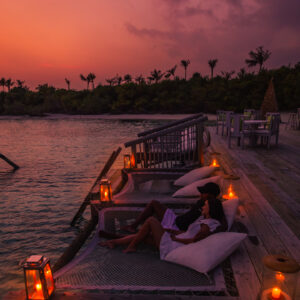 A couple relaxes on large cushions by a wooden deck, overlooking the water at sunset. Lanterns illuminate the scene, casting a warm glow. Nearby, a table and chairs are set up under the palm tree-lined shoreline, blending into the evening sky&#039;s gradient of purple and orange hues—truly embodying Soneva Exclusive Offers.