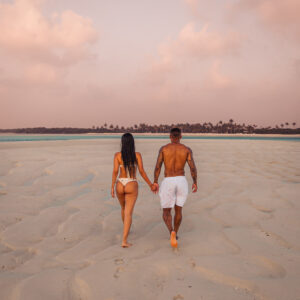 A man and woman holding hands while walking on a sandy beach at sunset. The man is shirtless and wearing white shorts, while the woman is in a white bikini. Their footprints trail behind them, reminiscent of Soneva Exclusive Offers that promise unforgettable romantic escapes amidst distant palm trees.