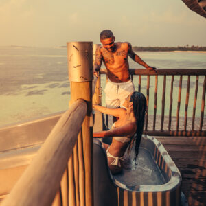 A man in swim trunks helps a woman in a white bikini out of an outdoor jacuzzi on a wooden deck at sunset, their laughter mingling with the waves. The woman’s long wet hair glistens under the golden glow, while his tattooed arm and chest create a picturesque moment. Experience similar scenes with Soneva Exclusive Offers.