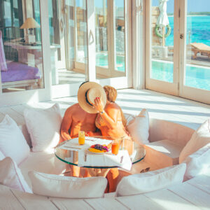 A couple sits in a cozy sunken seating area enjoying breakfast at a round glass table. They are sharing a kiss, with one person holding a straw hat that partially covers their faces. The scene, reminiscent of Soneva Exclusive Offers, overlooks a pool and beautiful ocean view through large glass doors.